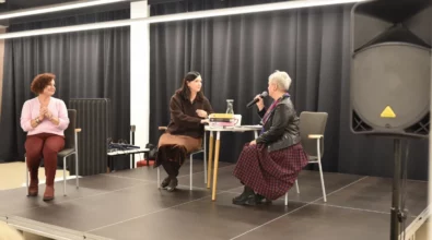 Joanna Kuciel-Frydryszak siedzi przy stoliku na scenie i rozmawia z prowadzącą spotkanie, która trzyma mikrofon. Na stoliku znajdują się książki autorki oraz karafka z wodą. Obok siedzi druga uczestniczka wydarzenia, która przysłuchuje się rozmowie.