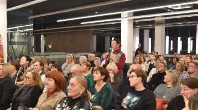 Uczestniczka spotkania stoi między rzędami krzeseł i trzyma mikrofon, zadając pytanie podczas dyskusji z publicznością. Wokół niej siedzą osoby uważnie słuchające wypowiedzi i obserwujące przebieg rozmowy. W wydarzeniu bierze udział liczna publiczność zgromadzona w sali Mediateki 800-lecia