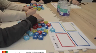 Uczestnicy warsztatów siedzą przy stole i układają kolorowe kostki z liczbami na macie edukacyjnej. Na blacie znajdują się materiały dydaktyczne z polami do sortowania oraz pojemniki z elementami do ćwiczeń. Zajęcia odbywają się w ramach projektu „MatSensoryka 4”, którego celem jest rozwijanie kompetencji matematycznych poprzez działania sensoryczne i praktyczne. Projekt został dofinansowany przez Fundację mBanku.