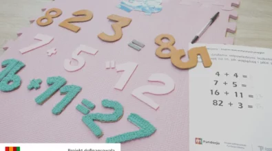 Na macie edukacyjnej ułożone są kolorowe cyfry i znaki matematyczne wykorzystywane podczas zajęć. Obok znajduje się karta pracy z przykładami działań matematycznych oraz długopis do zapisywania wyników. Materiały służą do nauki liczenia i rozwiązywania prostych zadań poprzez działanie i manipulowanie elementami. Zajęcia realizowane są w ramach projektu „MatSensoryka 4”, dofinansowanego przez Fundację mBanku.