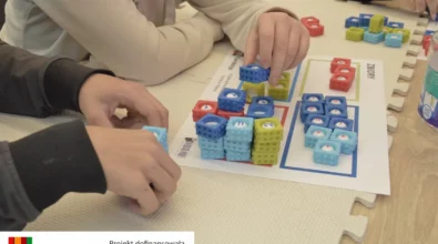 Dzieci układają kolorowe kostki z cyframi na planszy edukacyjnej, dopasowując je do wyznaczonych pól oznaczonych różnymi kolorami. Zadanie polega na porządkowaniu liczb i tworzeniu prostych działań matematycznych, co rozwija logiczne myślenie, koncentrację oraz umiejętność rozpoznawania liczb podczas zajęć w Mediatece 800-lecia realizowanych w ramach projektu „MatSensoryka 4”, dofinansowanego przez Fundację mBanku.