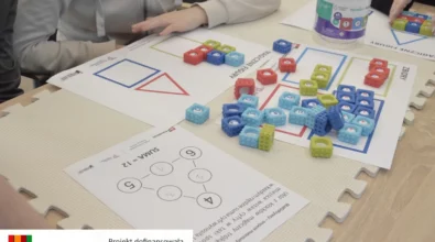 Uczestnicy warsztatów pracują przy stole, układając kolorowe kostki z liczbami na kartach pracy z zadaniami matematycznymi. Na macie edukacyjnej znajdują się plansze z figurami geometrycznymi oraz ćwiczeniami wymagającymi dopasowania liczb do odpowiednich pól. Zadania rozwijają umiejętność liczenia, logicznego myślenia i rozwiązywania prostych działań matematycznych poprzez manipulowanie elementami. Zajęcia realizowane są w ramach projektu „MatSensoryka 4”, dofinansowanego przez Fundację mBanku.