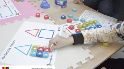 Uczestnik zajęć układa kolorowe kostki z liczbami na karcie pracy przedstawiającej figury geometryczne. Na macie edukacyjnej znajdują się również inne elementy wykorzystywane podczas ćwiczeń matematycznych. Zadanie polega na dopasowywaniu liczb i elementów do odpowiednich pól oraz figur. Zajęcia realizowane są w ramach projektu „MatSensoryka 4”, dofinansowanego przez Fundację mBanku.