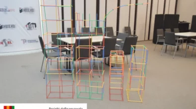 Na środku sali ustawione są wysokie konstrukcje z kolorowych rurek połączonych w geometryczne bryły. Powstałe struktury przypominają wieże i przestrzenne formy zbudowane podczas zajęć konstrukcyjnych. W tle widoczna jest przestrzeń Mediateki 800-lecia z ustawionymi stolikami i krzesłami oraz ścianka z logotypami instytucji. Konstrukcje powstały w trakcie zajęć realizowanych w ramach projektu „MatSensoryka 4”, dofinansowanego przez Fundację mBanku.