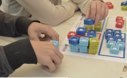 Dzieci układają kolorowe kostki z cyframi na planszy edukacyjnej, dopasowując je do wyznaczonych pól oznaczonych różnymi kolorami. Zadanie polega na porządkowaniu liczb i tworzeniu prostych działań matematycznych, co rozwija logiczne myślenie, koncentrację oraz umiejętność rozpoznawania liczb podczas zajęć w Mediatece 800-lecia realizowanych w ramach projektu „MatSensoryka 4”, dofinansowanego przez Fundację mBanku.