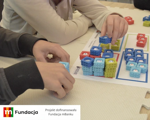 Dzieci układają kolorowe kostki z cyframi na planszy edukacyjnej, dopasowując je do wyznaczonych pól oznaczonych różnymi kolorami. Zadanie polega na porządkowaniu liczb i tworzeniu prostych działań matematycznych, co rozwija logiczne myślenie, koncentrację oraz umiejętność rozpoznawania liczb podczas zajęć w Mediatece 800-lecia realizowanych w ramach projektu „MatSensoryka 4”, dofinansowanego przez Fundację mBanku.