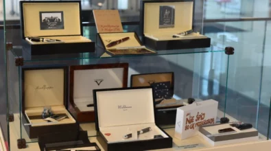 W szklanej gablocie ekspozycyjnej zaprezentowane są pióra wieczne umieszczone w eleganckich pudełkach i etui, ułożone na kilku poziomach wraz z kartami informacyjnymi. Zdjęcie przedstawia fragment wystawy prezentowanej w przestrzeni Miejskiej Biblioteki Publicznej.