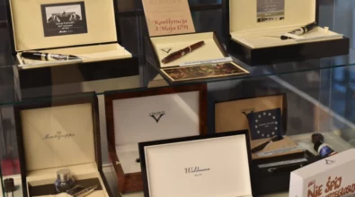 Na zdjęciu widoczna jest szklana gablota z ekskluzywnymi piórami wiecznymi umieszczonymi w eleganckich pudełkach. Na górnej półce znajdują się zestawy piór renomowanych marek, m.in. Montegrappa, ozdobione starannie wykonanymi detalami. Obok nich umieszczone są tabliczki z cytatami oraz zdjęciami wybitnych postaci, takich jak Maria Skłodowska-Curie i Władysław Reymont. Dolna półka zawiera kolejne pióra, w tym model marki Visconti w eleganckim drewnianym etui. W tle odbijają się sylwetki osób oglądających wystawę.