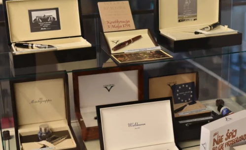 Na zdjęciu widoczna jest szklana gablota z ekskluzywnymi piórami wiecznymi umieszczonymi w eleganckich pudełkach. Na górnej półce znajdują się zestawy piór renomowanych marek, m.in. Montegrappa, ozdobione starannie wykonanymi detalami. Obok nich umieszczone są tabliczki z cytatami oraz zdjęciami wybitnych postaci, takich jak Maria Skłodowska-Curie i Władysław Reymont. Dolna półka zawiera kolejne pióra, w tym model marki Visconti w eleganckim drewnianym etui. W tle odbijają się sylwetki osób oglądających wystawę.