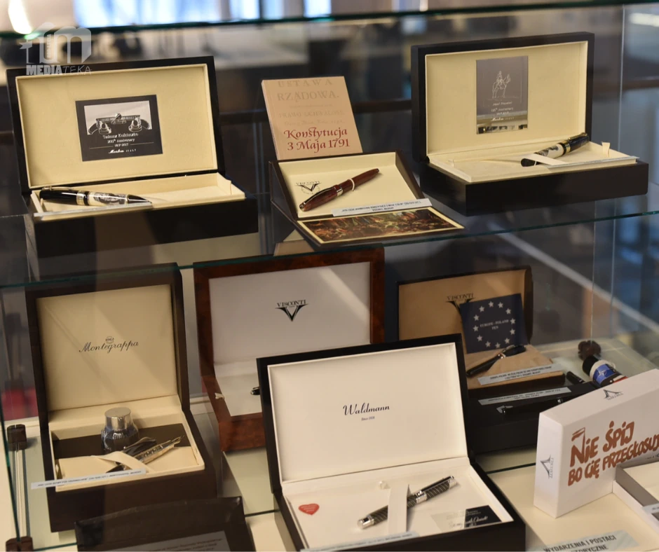 Na zdjęciu widoczna jest szklana gablota z ekskluzywnymi piórami wiecznymi umieszczonymi w eleganckich pudełkach. Na górnej półce znajdują się zestawy piór renomowanych marek, m.in. Montegrappa, ozdobione starannie wykonanymi detalami. Obok nich umieszczone są tabliczki z cytatami oraz zdjęciami wybitnych postaci, takich jak Maria Skłodowska-Curie i Władysław Reymont. Dolna półka zawiera kolejne pióra, w tym model marki Visconti w eleganckim drewnianym etui. W tle odbijają się sylwetki osób oglądających wystawę.