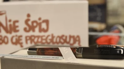 Na zdjęciu widoczne jest eleganckie pióro wieczne marki Visconti, umieszczone w otwartym pudełku wyłożonym jasnym materiałem. Na korpusie pióra znajduje się wygrawerowany napis „Święto Wolności 1989–2019”. W tle widoczna jest biała tabliczka z dużym, czerwonym napisem „Nie śpij, bo cię przegłosują”, co nawiązuje do hasła promującego obywatelskie zaangażowanie w wybory. Rozmyte elementy dalszego planu wskazują, że zdjęcie zostało wykonane w przestrzeni wystawowej.