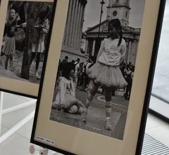 Oprawiona czarno-biała fotografia przedstawiająca dziewczynkę stojącą na ulicy, ubraną w jasną sukienkę, z innymi dziećmi widocznymi w tle miejskiej przestrzeni. Zdjęcie ukazuje fragment wystawy fotograficznej prezentowanej w Miejskiej Bibliotece Publicznej.
