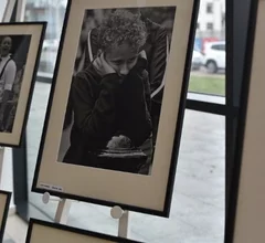 Oprawiona czarno-biała fotografia przedstawiająca młodą osobę pochyloną nad kartką lub notesem, skupioną na pisaniu lub rysowaniu, z widocznym tłem miejskim za przeszkloną ścianą. Zdjęcie ukazuje fragment wystawy fotograficznej prezentowanej w przestrzeni Miejskiej Biblioteki Publicznej.