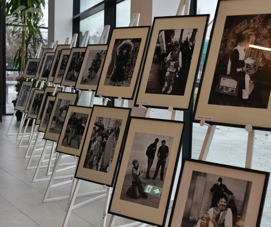 Na sztalugach ustawionych w długim rzędzie zaprezentowane są oprawione czarno-białe fotografie przedstawiające sceny z życia codziennego i portrety ludzi. Zdjęcie ukazuje fragment wystawy fotograficznej prezentowanej w przestrzeni Miejskiej Biblioteki Publicznej.