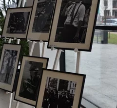 Na sztalugach ustawione są oprawione czarno-białe fotografie przedstawiające ludzi w przestrzeni miejskiej, uchwyconych w codziennych sytuacjach. Zdjęcie ukazuje fragment wystawy fotograficznej prezentowanej w przestrzeni Miejskiej Biblioteki Publicznej.