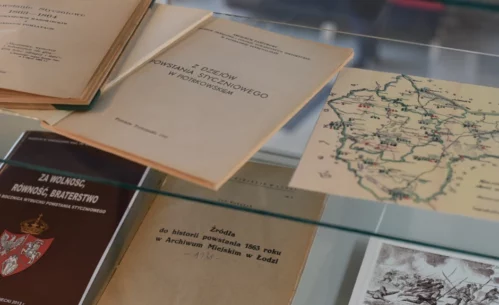 W szklanej gablocie wystawowej ułożone są archiwalne książki, broszury i materiały kartograficzne związane z historią XIX wieku. Zdjęcie przedstawia fragment wystawy historycznej prezentowanej w Miejskiej Bibliotece Publicznej im. Adama Próchnika.