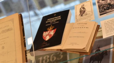 Na szklanej gablocie ekspozycyjnej wyeksponowane są stare książki i dokumenty związane z historią Polski, w tym publikacje z XIX wieku dotyczące okresu Powstania Styczniowego. Zdjęcie przedstawia fragment wystawy historycznej prezentowanej w Miejskiej Bibliotece Publicznej im. Adama Próchnika.