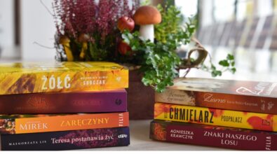 Na zdjęciu widoczne są stosy książek ułożone na stole, z wyraźnie widocznymi tytułami na grzbietach. W tle znajduje się jesienna dekoracja z wrzosu, zielonych roślin, jabłek i grzybów. Kompozycja utrzymana jest w ciepłej, naturalnej kolorystyce. Na dole grafiki umieszczono logotypy Narodowego Programu Rozwoju Czytelnictwa 2.0, Ministerstwa Kultury i Dziedzictwa Narodowego oraz Biblioteki Narodowej.