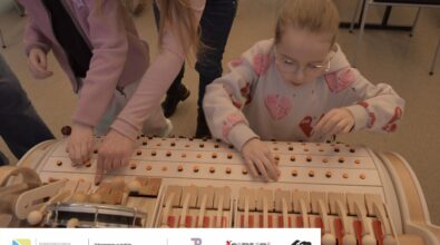 Na zdjęciu grupa osób zgromadzona przy dużym, drewnianym urządzeniu w kształcie walca. Urządzenie wyposażone jest w wiele rzędów pomarańczowych przycisków. Na urządzeniu ustawione są cymbałki, dodo i bębenek. Wszyscy wciskają pomarańczowe przyciski urządzenia. Zdjęcie MBP.