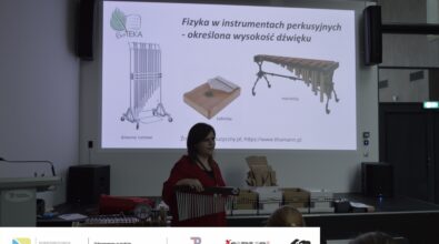 Zdjęcie przedstawiające kobietę w czerwonej koszuli. Stoi ona na tle prezentacji, na której widoczne są trzy instrumenty - dzwony rurowe, kalimba i marimba. Na podwyższeniu w tle stoją czarno-białe pudełka. Kobieta trzyma w dłoni instrument, który jest metalowymi rurkami o różnych długościach, ustawionych w kolejności od najdłuższej do najkrótszej. Zdjęcie: MBP.