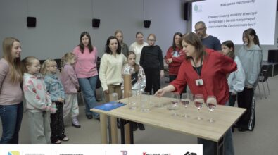 Na zdjęciu widać kobietę w czerwonej koszuli. Nachyla się ona nad stołem, na którym stoją przezroczyste naczynia wypełnione wodą. Obok stołu stoją różnej wysokości tekturowe rury. Na stół patrzy grupa osób - dorosłych wraz z dziećmi. W tle widać prezentację dotyczącą nietypowych instrumentów. Zdjęcie: MBP.