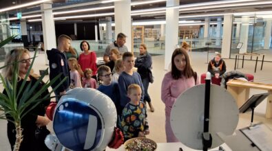 Zdjęcie przedstawia grupę ludzi stojących przedem do zdjęcia. Znajdują się przed stolikiem z kołem fortuny, zapewne czekają na swoją kolej w konkursie. Obok stolika stoi duży balon w kształcie astronauty. W tle widać teren biblioteki. Zdjęcie: MBP.