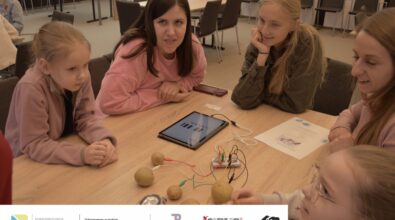 Zdjęcie przedstawia dwie dziewczynki i trzy kobiety siedzące przy stole. Na stole leżą ziemniaki połączone kolorowymi przewodami z płytką elektroniczną i tabletem. Na tablecie wyświetla się klawiatura pianina. Obok leży instrukcja. Zdjęcie MBP.