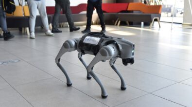 Zdjęcie przedstawia robota psa. Jest szary, na tułowiu ma czarną otoczkę. Stoi w dużej przestrzeni, w tle widać oglądających go ludzi. Zdjęcie: MBP.