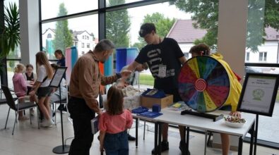 Zdjęcie przedstawia mężczyznę z dzieckiem stojących obok stołu z kołem fortuny. Rozmawiają z nastolatkiem w czarnej koszulce, który ma na szyi identyfikator. Po prawej stronie obok stołu stoi stojak z informacją odbywającym się konkursie z kołem fortuny. W tle widać drugą grupę nastolatków przy stoliku. Zdjęcie: MBP.