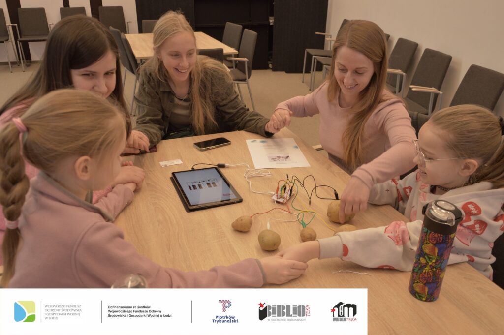 Zdjęcie przedstawia dwie dziewczynki i trzy kobiety siedzące przy stole. Wszystkie osoby trzymają się za ręce i się uśmiechają. Na stole leżą ziemniaki połączone kolorowymi przewodami z płytką elektroniczną i tabletem. Na tablecie wyświetla się klawiatura pianina. Obok leży instrukcja. Zdjęcie MBP.