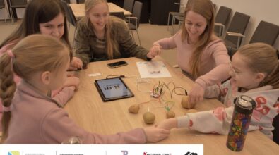 Zdjęcie przedstawia dwie dziewczynki i trzy kobiety siedzące przy stole. Wszystkie osoby trzymają się za ręce i się uśmiechają. Na stole leżą ziemniaki połączone kolorowymi przewodami z płytką elektroniczną i tabletem. Na tablecie wyświetla się klawiatura pianina. Obok leży instrukcja. Zdjęcie MBP.