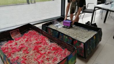 Zdjęcie przedstawiające dwa duże pudełka. Ich boki pomalowane są na różne kolory. Wnętrza wypełniono piaskiem kinetycznym. W pudełku bliżej osoby fotografującej piasek ma kolor czerwony i znajdują się w nim elementy z czerwonej bibuły, a w drugim pudełku piasek ma bardzo jasny kolor. Na piasku w obu pudełkach leżą podpisane grafiki marsa (czerwony) oraz księżyca (jasny). Na jasny piasek nastolatek stawia prototyp łazika kosmicznego, będący niebiesko-fioletowym pudełkiem z trzema widocznymi kołami. W tle widać chodnik oraz biały stół. Zdjęcie: MBP.