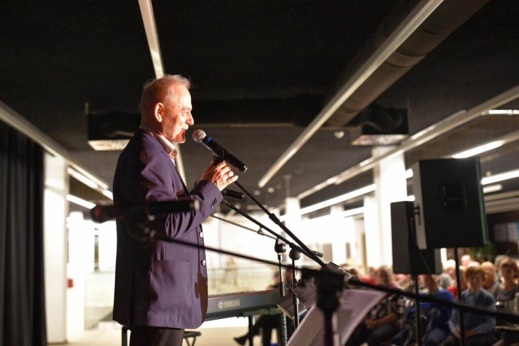 Na zdjęciu widoczny jest mężczyzna stojący na scenie i przemawiający do mikrofonu. Ubrany jest w elegancką marynarkę, a jego sylwetka oświetlona jest światłem scenicznym, które podkreśla profil twarzy. Przed nim znajdują się statywy z mikrofonami oraz pulpit z nutami lub notatkami, a w tle widoczna jest klawiatura oraz elementy nagłośnienia. W głębi kadru można dostrzec siedzącą publiczność, skupioną na wystąpieniu, co wskazuje na oficjalny charakter wydarzenia odbywającego się w nowoczesnej przestrzeni Mediateki.