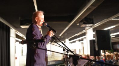 Na zdjęciu widoczny jest mężczyzna stojący na scenie i przemawiający do mikrofonu. Ubrany jest w elegancką marynarkę, a jego sylwetka oświetlona jest światłem scenicznym, które podkreśla profil twarzy. Przed nim znajdują się statywy z mikrofonami oraz pulpit z nutami lub notatkami, a w tle widoczna jest klawiatura oraz elementy nagłośnienia. W głębi kadru można dostrzec siedzącą publiczność, skupioną na wystąpieniu, co wskazuje na oficjalny charakter wydarzenia odbywającego się w nowoczesnej przestrzeni Mediateki.