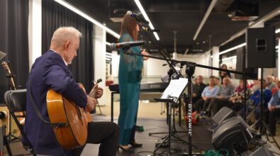 Na zdjęciu widoczna jest scena koncertowa w nowoczesnej sali. Na pierwszym planie po lewej stronie siedzi mężczyzna grający na gitarze akustycznej, ubrany w marynarkę, skupiony na instrumencie. W centralnej części kadru stoi kobieta przy mikrofonie, ubrana w długi, zielono-turkusowy strój, zwrócona w stronę publiczności. Przed nią znajduje się pulpit z nutami oraz klawiatura. Na scenie widoczne są statywy mikrofonowe, kable i nagłośnienie. W tle, po prawej stronie, siedzi publiczność uważnie słuchająca występu. Całość oddaje atmosferę kameralnego koncertu lub wydarzenia kulturalnego.