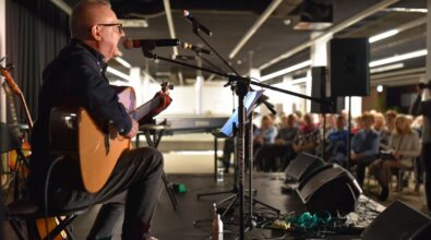 Na zdjęciu widoczny jest mężczyzna siedzący na krześle na scenie, grający na gitarze akustycznej i śpiewający do mikrofonu umieszczonego na statywie. Ma na sobie ciemną koszulę, spodnie oraz jasne buty, a na nosie okulary. Przed nim znajduje się pulpit z nutami, a wokół ustawione są mikrofony, monitory odsłuchowe, kable oraz elementy nagłośnienia. W tle widać zgromadzoną publiczność siedzącą na krzesłach, uważnie słuchającą występu, a także instrumenty muzyczne, w tym keyboard. Całość odbywa się w nowoczesnej, przestronnej sali, oświetlonej ciepłym światłem scenicznym, co podkreśla kameralny charakter koncertu.