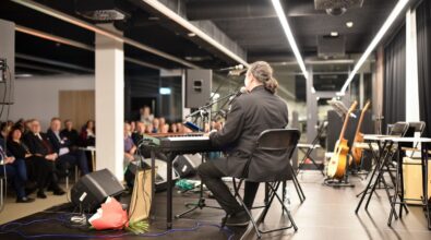 Na zdjęciu widoczny jest muzyk siedzący tyłem do aparatu przy elektronicznej klawiaturze na scenie, grający i śpiewający do mikrofonu. Artysta ubrany jest w ciemny strój i siedzi na składanym krześle, a obok niego na podłodze leży bukiet kwiatów oraz przewody nagłośnieniowe. Na scenie ustawione są statywy z mikrofonami, głośniki oraz instrumenty, w tym gitary stojące na stojakach i dodatkowa klawiatura. W tle widać zgromadzoną publiczność siedzącą na krzesłach, uważnie słuchającą koncertu w nowoczesnej, dobrze oświetlonej przestrzeni Mediateki.