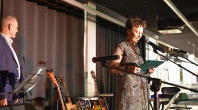 Na zdjęciu widoczna jest kobieta stojąca na scenie i czytająca tekst z zielonej teczki, trzymając ją obiema rękami. Przemawia do mikrofonu ustawionego na statywie, a jej twarz jest oświetlona światłem scenicznym. Ubrana jest w elegancką, jasną sukienkę z długimi rękawami. Po lewej stronie kadru widać mężczyznę stojącego przy pulpicie z nutami, a w tle ustawione są gitary na stojakach oraz instrumenty klawiszowe. Całość odbywa się w nowoczesnej sali koncertowej z czarną kurtyną i oświetleniem scenicznym, co podkreśla oficjalny i uroczysty charakter wydarzenia.