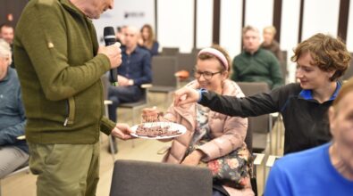 Mężczyzna trzyma mikrofon i podaje uczestnikom spotkania talerz z kawałkami czekolady na wykałaczkach. Siedząca kobieta z uśmiechem sięga po poczęstunek, a obok niej kolejna osoba wybiera czekoladę. W tle widoczni są pozostali uczestnicy wydarzenia siedzący na krzesłach. Scena przedstawia swobodną i życzliwą atmosferę spotkania w sali Mediateki.