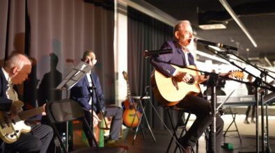 Na zdjęciu widoczny jest koncert odbywający się na scenie w nowoczesnej przestrzeni Mediateki. Na pierwszym planie starszy mężczyzna siedzi na wysokim krześle i śpiewa do mikrofonu, jednocześnie grając na gitarze akustycznej; ubrany jest w elegancką marynarkę i koszulę. Po lewej stronie kadru inny muzyk gra na gitarze basowej, siedząc na krześle i pochylając się nad instrumentem. W tle widoczny jest kolejny wykonawca siedzący przy instrumencie perkusyjnym typu cajón oraz stojąca gitara oparta o statyw. Na scenie znajdują się mikrofony na statywach, pulpit z nutami oraz klawiatura elektroniczna, a całość oświetlona jest światłem scenicznym. Atmosfera zdjęcia sugeruje kameralny, nastrojowy koncert z udziałem kilku instrumentalistów.