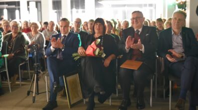 Na zdjęciu widoczna jest publiczność siedząca w pierwszym rzędzie podczas wydarzenia kulturalnego. Kilka osób, kobiety i mężczyźni w eleganckich strojach, bije brawo, wyrażając uznanie dla występu. W centrum kadru znajduje się kobieta trzymająca bukiet kwiatów, obok niej mężczyzna z teczką oraz inny mężczyzna w garniturze, również klaszczący. Na podłodze przy jednym z krzeseł widoczna jest oprawiona grafika, a z przodu ustawiony jest niewielki statyw z kamerą lub aparatem. W tle widać liczną publiczność siedzącą na krzesłach w jasnej, nowoczesnej sali, oświetlonej światłem scenicznym, co podkreśla uroczysty charakter wydarzenia.