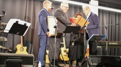 Na zdjęciu widoczna jest uroczysta scena na estradzie, podczas której kilku mężczyzn w eleganckich garniturach wręcza wyróżnienie artyście. Jeden z mężczyzn podaje dokument w ozdobnej teczce, inny trzyma oprawiony dyplom, a w tle widać osobę z bukietem kwiatów. Na scenie ustawione są statywy z mikrofonami, pulpity nutowe oraz instrumenty muzyczne, w tym gitary i klawiatura. Całość odbywa się w nowoczesnej sali Mediateki, a atmosfera wydarzenia ma charakter oficjalny i podniosły, podkreślający rangę uhonorowania oraz znaczenie wydarzenia kulturalnego.