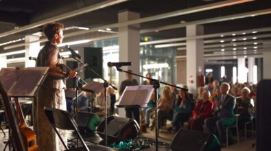 Na zdjęciu widoczna jest artystka stojąca na scenie i przemawiająca do zgromadzonej publiczności. Kobieta trzyma w dłoniach kartkę i stoi przy mikrofonie, a przed nią ustawione są statywy z nutami, głośniki oraz instrumenty muzyczne, w tym gitara oparta o krzesło. W tle widać licznie zgromadzoną publiczność siedzącą na krzesłach, która z uwagą słucha wystąpienia, część osób bije brawo. Wydarzenie odbywa się w nowoczesnej, jasnej przestrzeni Mediateki, a atmosfera ma charakter uroczysty i kulturalny.