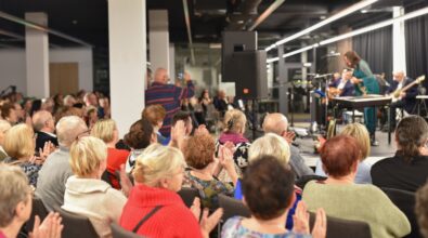 Na zdjęciu widoczna jest publiczność zgromadzona w nowoczesnej sali koncertowej, siedząca na krzesłach i głośno bijąca brawo po zakończonym występie. W pierwszym planie widać plecy i profile uczestników, głównie osób starszych, ubranych w swobodne, kolorowe stroje. W centralnej części kadru stoi mężczyzna nagrywający wydarzenie telefonem komórkowym. W tle, na podwyższonej scenie, znajdują się artyści z instrumentami muzycznymi, mikrofonami i nagłośnieniem, a całość oświetlona jest światłem scenicznym. Zdjęcie oddaje atmosferę uznania, zaangażowania i wspólnego przeżywania wydarzenia kulturalnego.