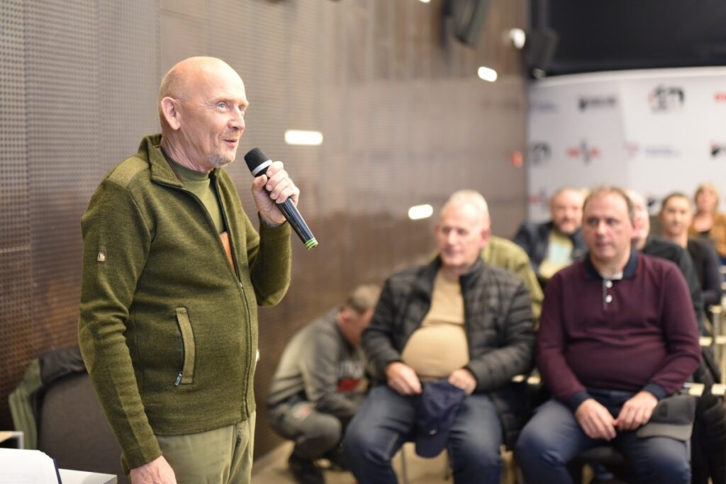 Mężczyzna stoi w sali Mediateki i przemawia do publiczności, trzymając mikrofon w dłoni. Za nim widoczni są uczestnicy spotkania siedzący na krzesłach i uważnie słuchający wystąpienia.