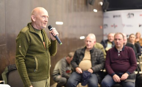 Mężczyzna stoi w sali Mediateki i przemawia do publiczności, trzymając mikrofon w dłoni. Za nim widoczni są uczestnicy spotkania siedzący na krzesłach i uważnie słuchający wystąpienia.