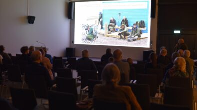 Uczestnicy spotkania siedzą na widowni i oglądają prezentację wyświetlaną na dużym ekranie. Na slajdzie widoczna jest fotografia grupy osób podczas wyprawy terenowej w plenerze. Prowadzący stoi z boku sali i komentuje prezentowane materiały, trzymając mikrofon. Zdjęcie oddaje skupioną atmosferę wykładu i zaangażowanie publiczności.