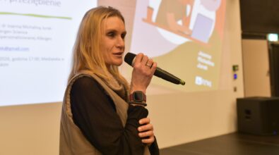 Kobieta przemawiająca do mikrofonu podczas prezentacji, w tle slajd z tekstem po polsku.