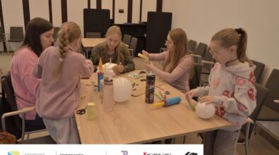 Na zdjęciu dwie dziewczynki i trzy kobiety zgromadzone wokół stołu. Na stole przybory plastyczne, styropian, balony i pompki do balonów. Jedna z kobiet tworzy jakąś konstrukcję ze styropianu i drewnianych patyczków. Druga klei coś żółtą taśmą. Trzecia kobieta pracuje nad czymś z dziewczynką, która jest odwrócona tyłem do obiektywu. Druga dziewczynka trzyma w dłoniach biały balonik. Zdjęcie MBP.