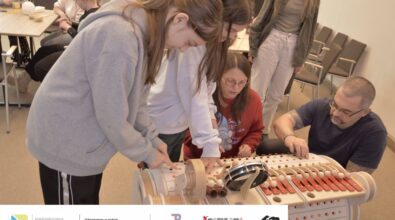 Na zdjęciu grupa osób zgromadzona przy dużym, drewnianym urządzeniu w kształcie walca. Urządzenie wyposażone jest w wiele rzędów pomarańczowych przycisków. Na urządzeniu ustawione są cymbałki, dodo i bębenek. Wszyscy wciskają pomarańczowe przyciski urządzenia. W tle inne osoby siedzące przy stolikach. Na stolikach balony i rolki po papierze. Zdjęcie MBP.